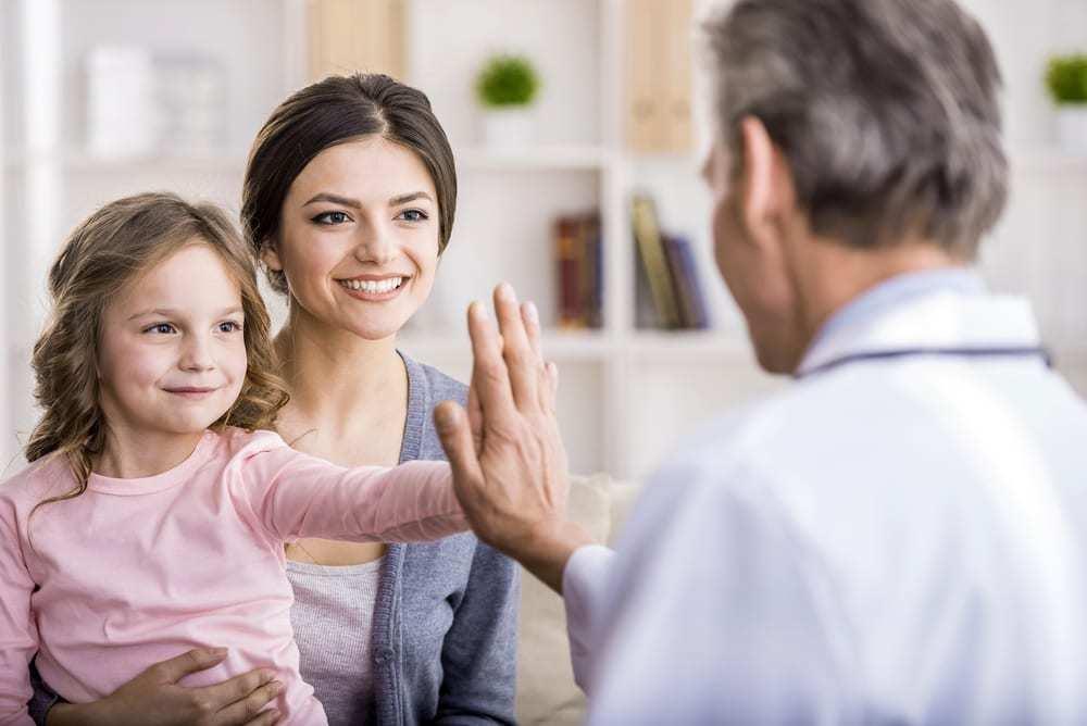 a dentist high-fiving a young child during a consultation, with the child's mother smiling in the background.