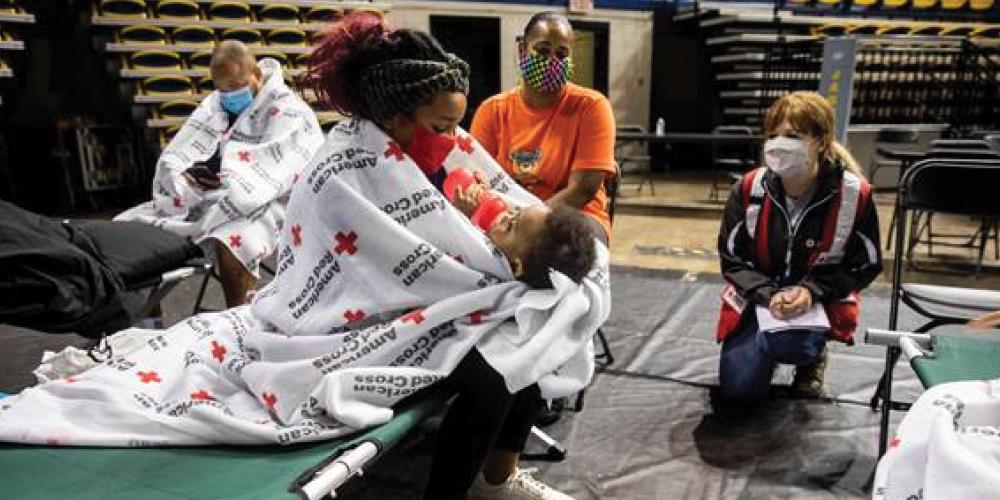 A woman sits on a cot in a gym, wrapped in Red Cross blankets while feeding her baby