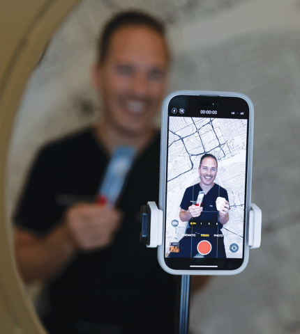 A dentist sitting behind a phone on a tripod showing him on the screen recording a video